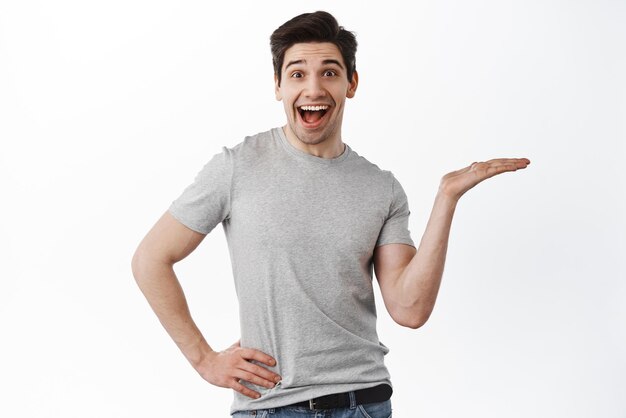 Excited funny guy holding on palm display your product in hand and smiling amazed showing object empty copy space standing over white background