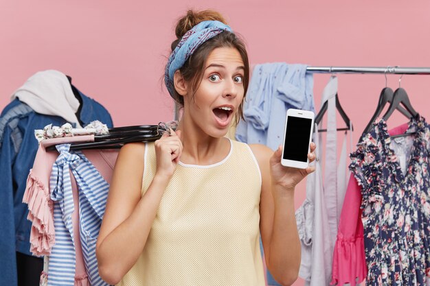 Excited funny woman shopaholic holding hangers with summer clothes to try on and demonstrating smartphone