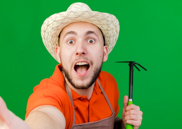 Excited gardener man wearing gardening hat holds hoe rake isolated