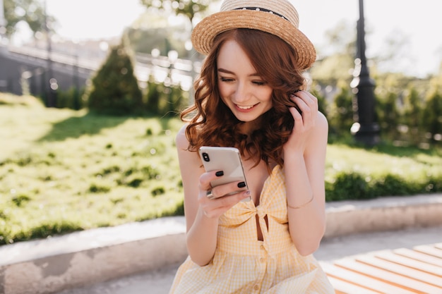 Excited ginger girl in hat texting message with smile. Outdoor portrait of sensual red-haired lady posing with her smartphone in park.