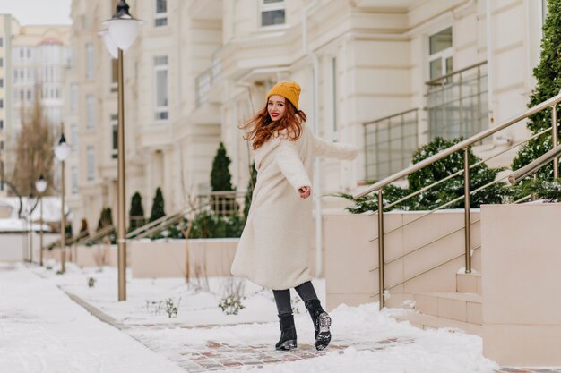雪道を歩きながら肩越しに見ている興奮した生姜少女。白いコートを着た魅力的な赤毛の女性の屋外ショット。