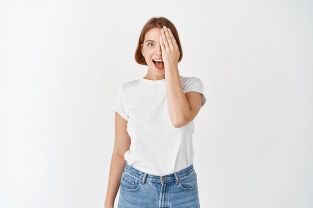 Excited girl cover half of face and eye, gasping amazed and looking at something awesome, standing in t-shirt and jeans on white wall