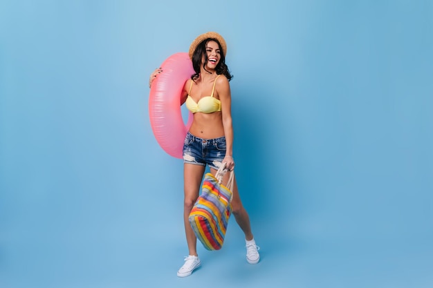 水泳サークルを保持しているデニムショートパンツの興奮した女の子青い背景で踊る麦わら帽子で官能的な日焼けした女性のスタジオショット