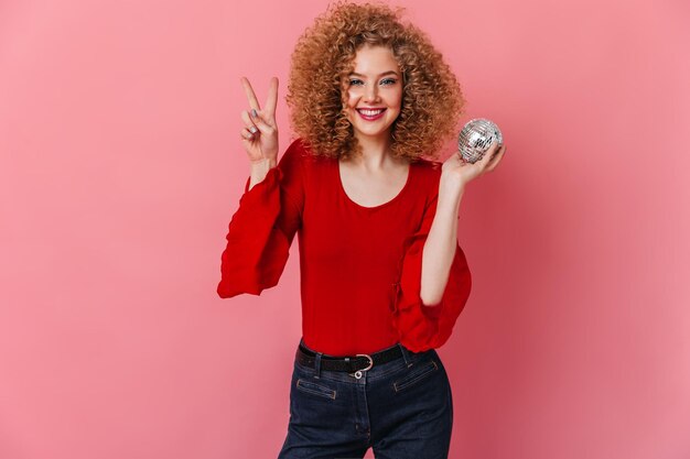 Excited girl dressed in red blouse and jeans posing with disco ball and showing sign of peace on pink background