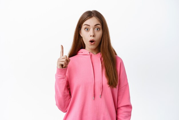 Excited girl gasping pointing finger up at copy space look impressed talking about advertisement standing over white background