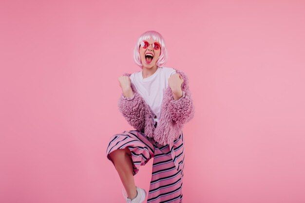 Excited girl in glamorous pink clothes expressing happiness Studio portrait of trendy shorthaired lady wears fluffy jacket and sunglasses