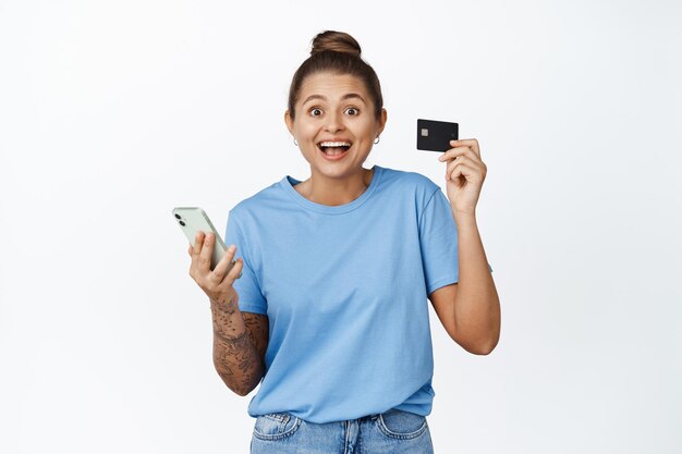 クレジットカードと携帯電話を持って、銀行のアプリで支払い、スマートフォンでオンラインショッピング、白い背景に立って興奮している女の子