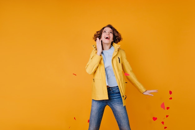 Excited girl in jeans and autumn coat jumping and throwing out paper hearts. Romantic active woman celebrating valentine's day in studio.