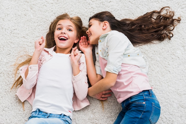 Excited girl listening to friend whispering to her ear lying on carpet