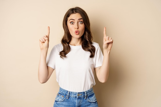 Excited girl looking interested pointing fingers up showing advertisement standing in tshirt over be...