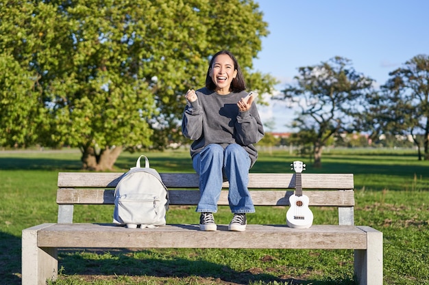 Foto gratuita la ragazza eccitata guarda lo smartphone e celebra le vittorie sul cellulare si siede con l'ukulele e lo zaino