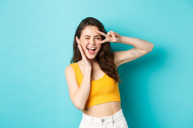 Excited girl screaming from joy, ready for summer holidays, showing v-sign and smiling happy, standing happy against blue background. Copy space