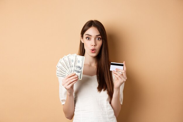 Excited girl showing dollar bills and plastic credit card saying wow with amazed face standing on be...