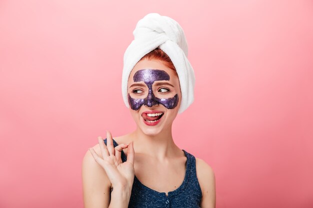 Возбужденная девушка показывает знак хорошо во время курортного лечения. Съемка студии счастливой молодой женщины с полотенцем и лицевой маской изолированные на розовой предпосылке.