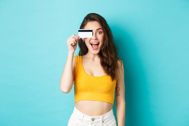 Excited girl in summer cropped top, look amazed at camera, shopping with credit card, standing over blue background.
