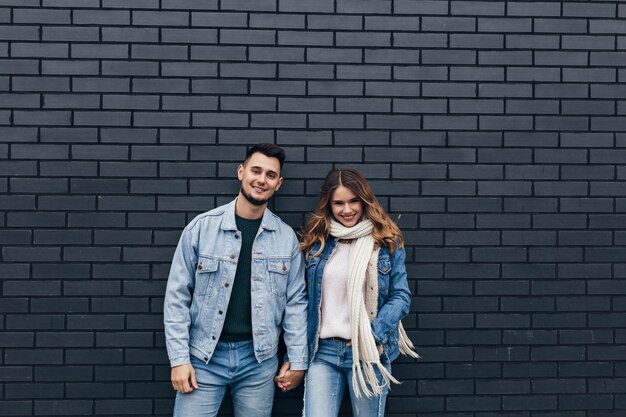 Excited girl in trendy denim outfit holding hands with boyfriend. Smiling loving couple standing together on bricked wall.