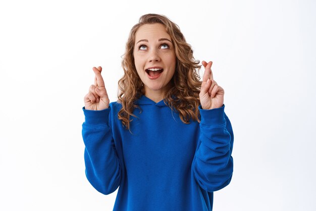 Excited girl willing to win, smiling and looking up with fingers crossed, making wish, waiting for lucky chance, praying, standing over white wall