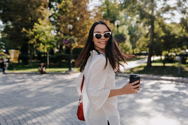 サングラスに白いシャツを着て素敵な笑顔で興奮した女の子がコーヒーを持って街の日当たりの良い緑豊かな公園を歩いている間カメラで振り返る