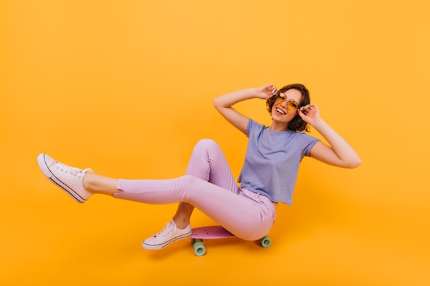 Excited girl in yellow-colored glasses sitting on longboard with cheerful smile. Jocund european lady with tattoo expressing positive emotions