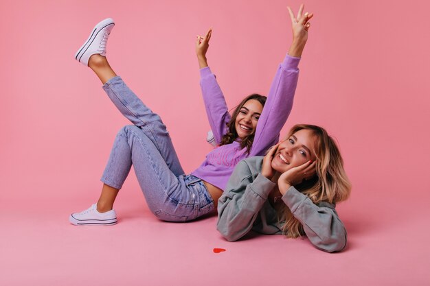 Excited girls in casual spring outfit posing on the floor. Positive best friends fooling around on pastel.