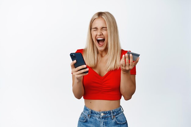 白い背景の上に立っているショッピングアプリでのオンライン販売に驚かされたスマートフォンとクレジットカードを保持しながら興奮した魅力的な女の子が悲鳴を上げる