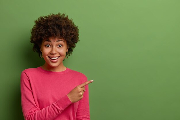 Excited good looking woman with Afro hair points finger right, sees good offer, suggests click link or follow page to find out information, has satisfied happy expression, dressed in casual clothes