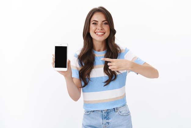 Взволнованная великолепная женщина объясняет функцию смартфона, представляет фотографии с летних каникул, радостно улыбается, указывая на экран мобильного телефона, подставку на белом фоне, продвигает онлайн-приложение