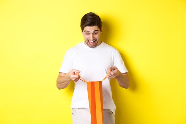 Excited guy open bag with gift looking inside with amazement and happy face standing against yellow ...