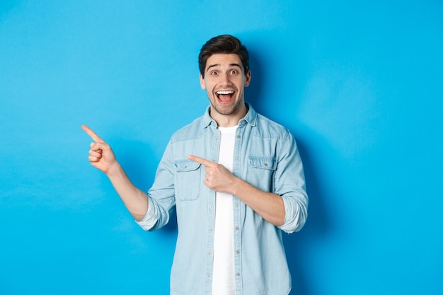 Возбужденный красивый 25-летний мужчина с бородой, указывая пальцами влево и изумленно улыбаясь, стоит на синем фоне
