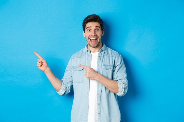 Excited handsome 25s years man with beard, pointing fingers left and smiling amazed, standing against blue background.