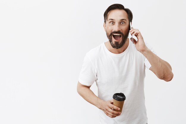 Excited handsome adult man drinking coffee and talking on smartphone