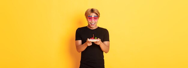 Excited handsome asian guy in party glasses looking hopeful at birthday cake as celebrating bday mak