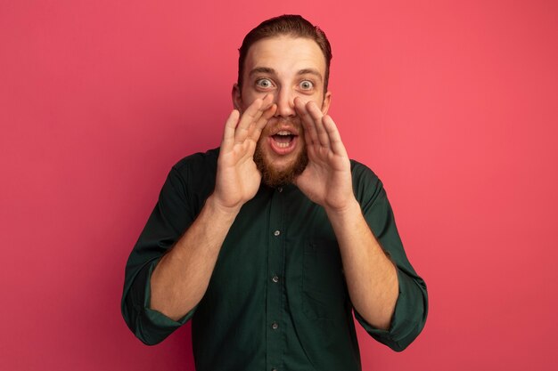 Excited handsome blonde man holds hands close to mouth isolated on pink wall