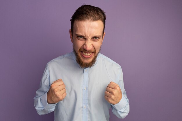 Excited handsome blonde man keeps fists looking at front isolated on purple wall