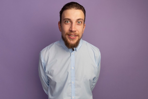 Excited handsome blonde man looking at front isolated on purple wall