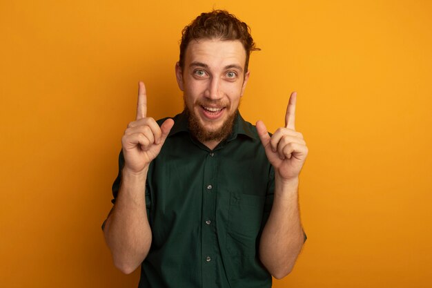 Excited handsome blonde man points up with two hands isolated on orange wall