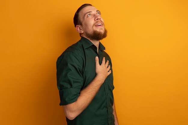 Excited handsome blonde man puts hand on chest and looks up isolated on orange wall