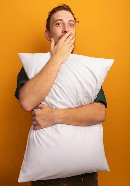 Excited handsome blonde man puts hand on mouth and holds pillow isolated on orange wall