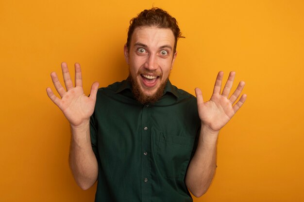 Excited handsome blonde man stands with raised hands isolated on orange wall