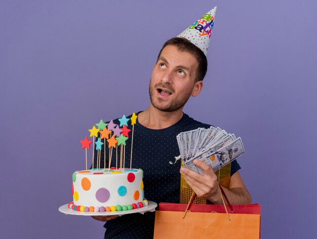 Excited handsome caucasian man wearing birthday cap holds birthday cake paper shopping bag gift box and money looking at side isolated on purple background with copy space