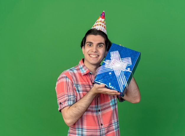 Excited handsome caucasian man wearing birthday cap holds gift box 