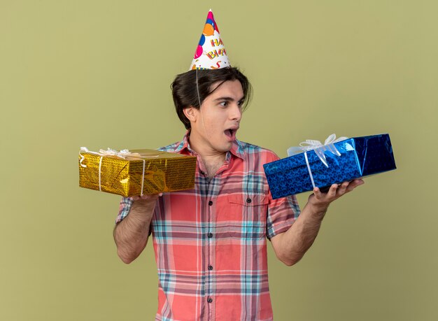 Excited handsome caucasian man wearing birthday cap holds and looks at gift boxes 