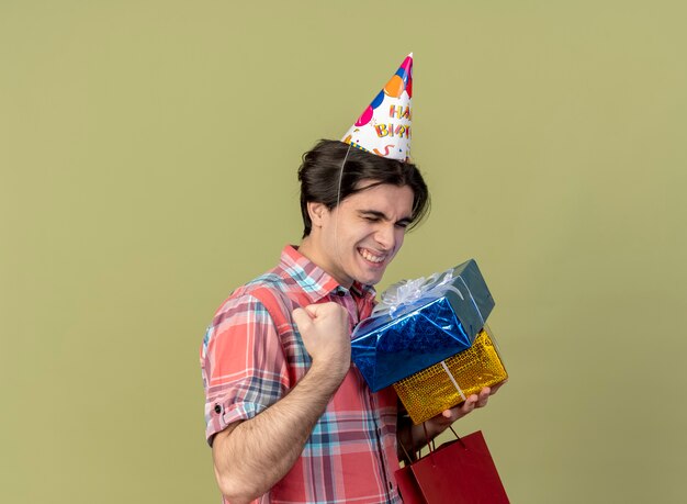 Excited handsome caucasian man wearing birthday cap keeps fist holds gift boxes and paper shopping bag 
