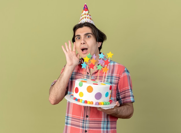 Free photo excited handsome caucasian man wearing birthday cap stands with raised hand and holds birthday cake