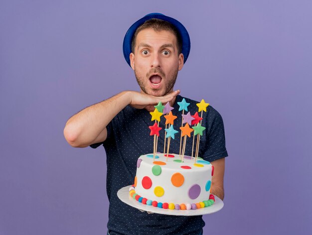 excited handsome caucasian man wearing blue hat keeps hand under chin and holds birthday cake isolated on purple background with copy space