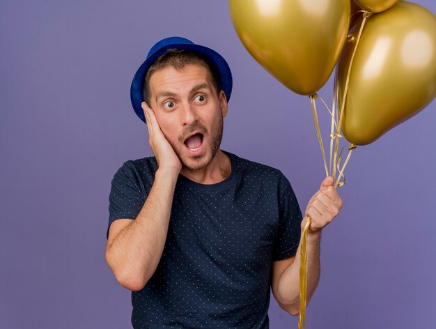 Excited handsome caucasian man wearing blue hat puts hand on face holds helium balloons isolated on purple background with copy space