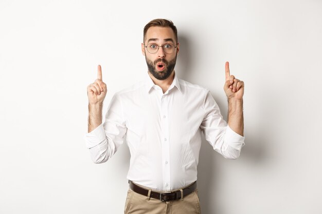 Excited handsome guy in glasses pointing fingers up, showing information banner, standing  white