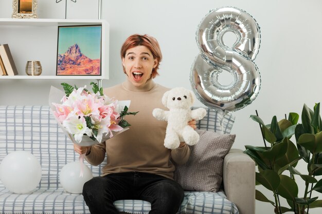 Excited handsome guy on happy women day holding bouquet with teddy bear sitting on sofa in living room