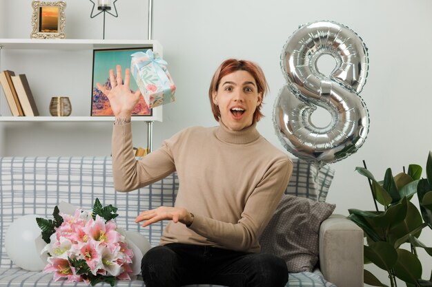 Excited handsome guy on happy women day toss a present sitting on sofa in living room