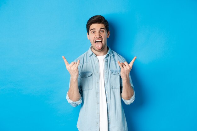 Excited handsome guy having fun, showing rock and roll sign and sticking tongue, standing against blue background.